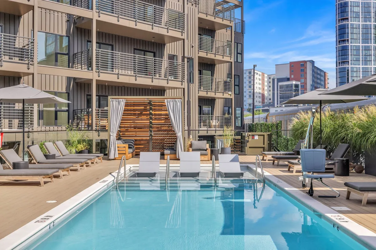 An inviting swimming pool area with lounge chairs, umbrellas, and the building's modern facade visible in the background. The pool deck looks ready for both morning workouts and after-hours relaxation, capturing the atmosphere of leisure and wellness.