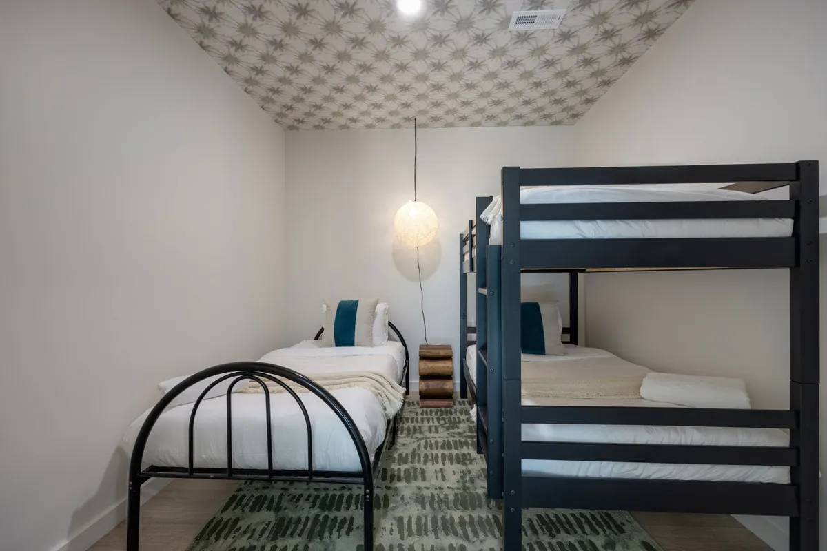 A playful and cozy second bedroom featuring a bunk bed and a single bed, illuminated by a modern pendant light and resting on a decorative rug. The space is inviting for families or groups, offering flexibility and comfort.