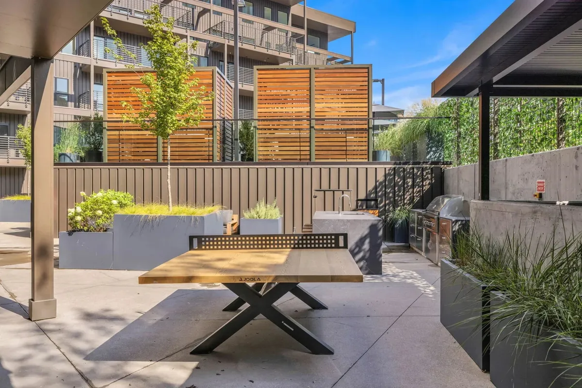 An exterior communal area featuring an outdoor dining table, modern grill station, wooden privacy screens, and vibrant planter boxes. The space is sunlit and inviting, designed for group meals and social gatherings.
