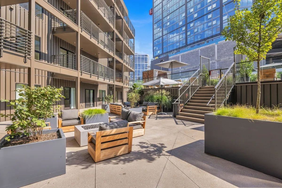 A stylish outdoor lounge area with comfortable seating, potted plants, a canopy umbrella, and a staircase leading to another level. The setting is ideal for relaxing, hosting small gatherings, or enjoying fresh air with friends.