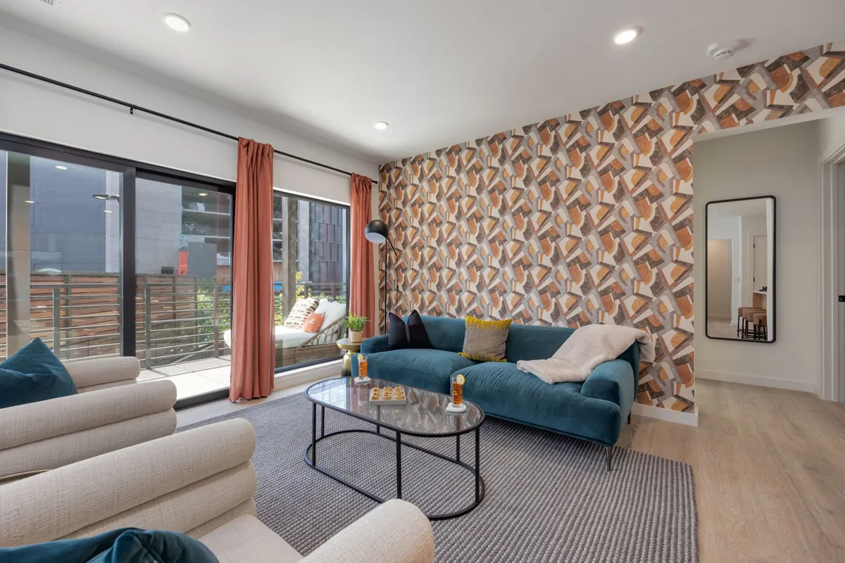 A sunlit living room with a comfortable teal sofa, a colorful accent wall, and large windows that invite abundant natural light. The space is set for socializing, relaxing, or hosting casual gatherings in a stylish, bohemian-inspired setting.