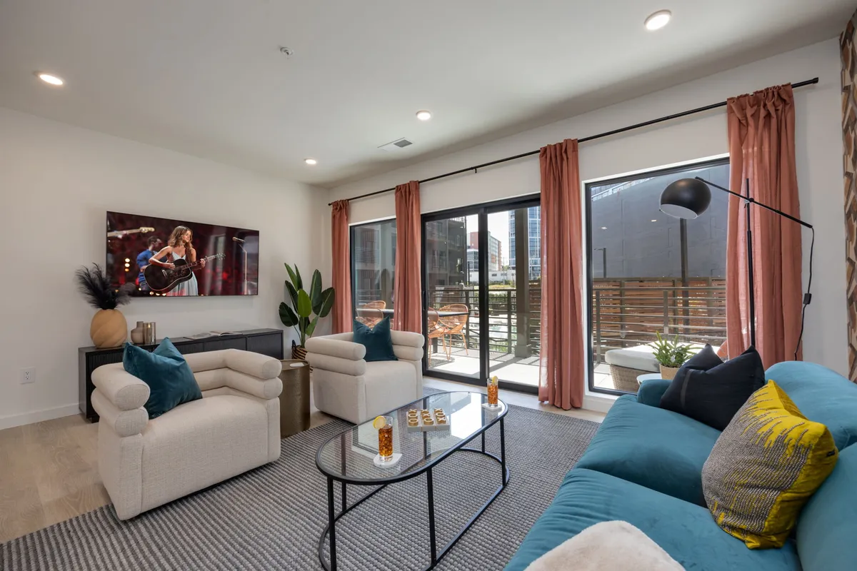 The image showcases a stylish, boho-inspired living room with a plush sofa, chic coffee table, large TV, and expansive windows letting in natural light. The space exudes comfort and sophistication, perfect for relaxing or entertaining, with a glimpse of the balcony inviting guests to enjoy their morning coffee.