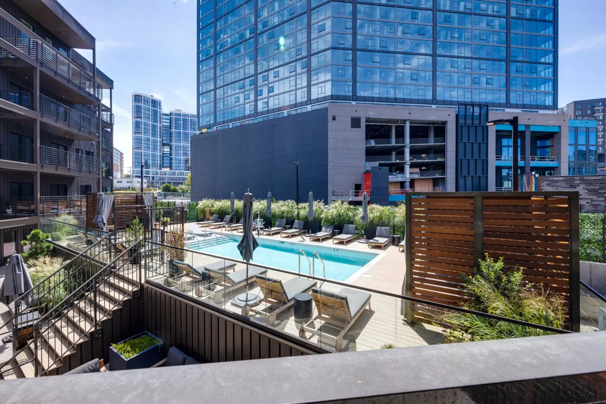 A vibrant, resort-style swimming pool surrounded by lounge chairs, landscaped green spaces, and a modern building facade. The setting is inviting, with ample room for sunbathing, swimming, and social gatherings by the water.