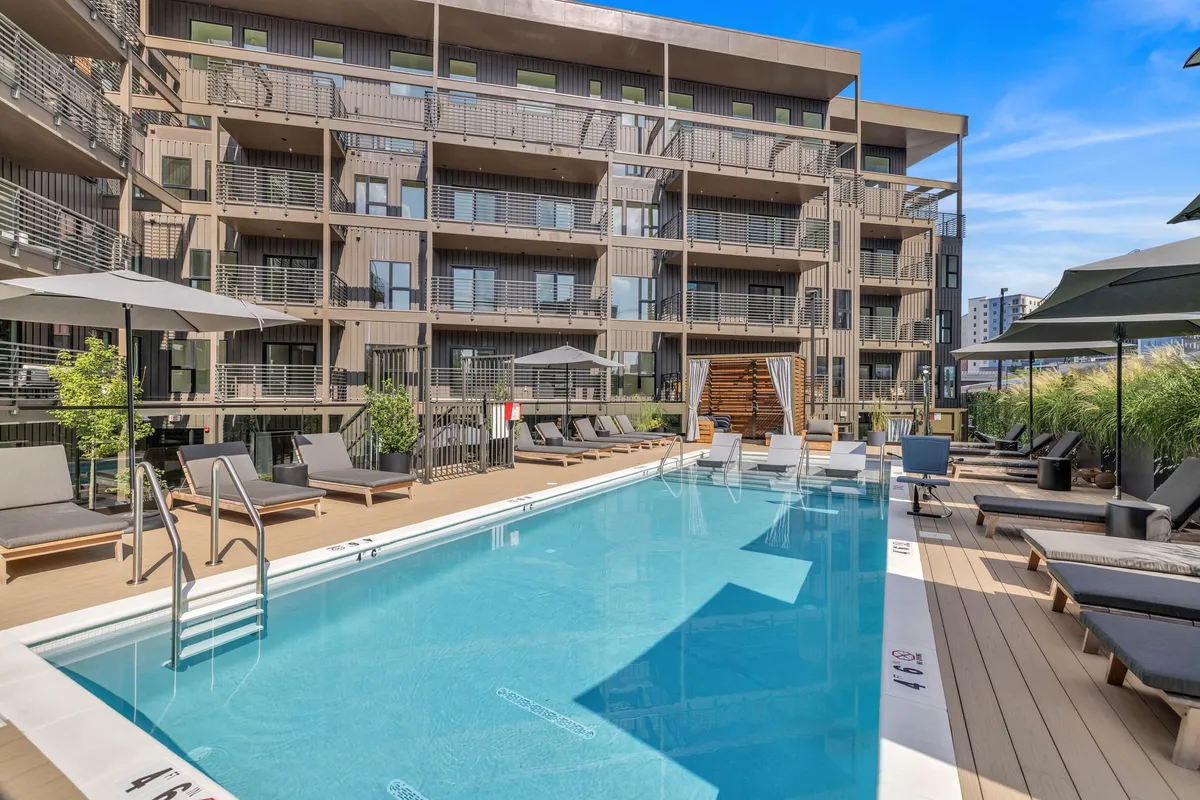 The image shows a resort-style swimming pool surrounded by lounge chairs and umbrellas, with the building’s facade rising behind. The atmosphere is relaxed and social, and the elevated perspective hints at the views available from above.