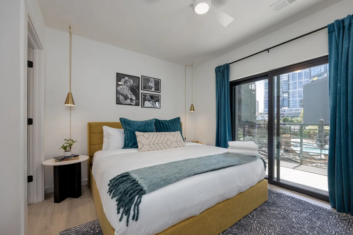 A stylish bedroom with a bed featuring decorative pillows and a blue throw blanket. Large windows allow natural light to flood the room, and framed photographs provide a personal touch. The windows offer a view of the city skyline, enhancing the sense of retreat and tranquility.
