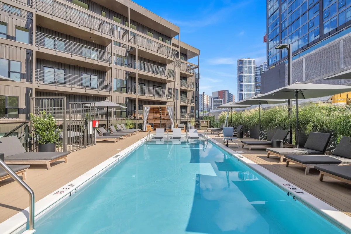 A modern outdoor swimming pool is surrounded by comfortable lounge chairs and set against the striking facade of a contemporary apartment building. The area is sunlit, inviting, and designed for both relaxation and socializing, with ample space for groups and poolside gatherings.