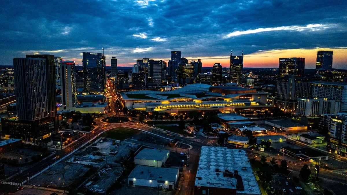 A dramatic city skyline at night with illuminated buildings, lit roadways, and a nearby park area. The image radiates energy and captures the pulse of Nashville after dark, hinting at both nightlife and city events.