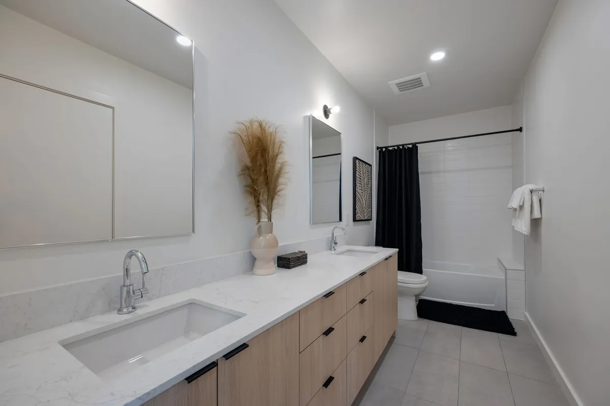 A contemporary bathroom with double sinks and mirrors, a decorative plant, and a black shower curtain. The room exudes a calming, spa-like atmosphere ideal for relaxation and self-care routines.